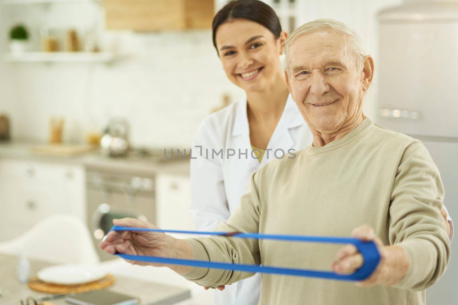 Happy senior citizen using fitness rubber band with female doctor in the background at home