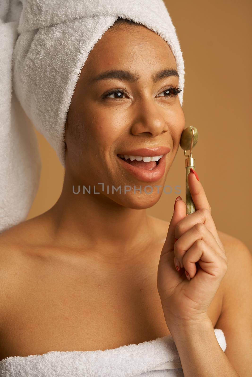 Portrait of happy young woman after shower smiling while using jade roller for massaging her face isolated over beige background. Skincare concept