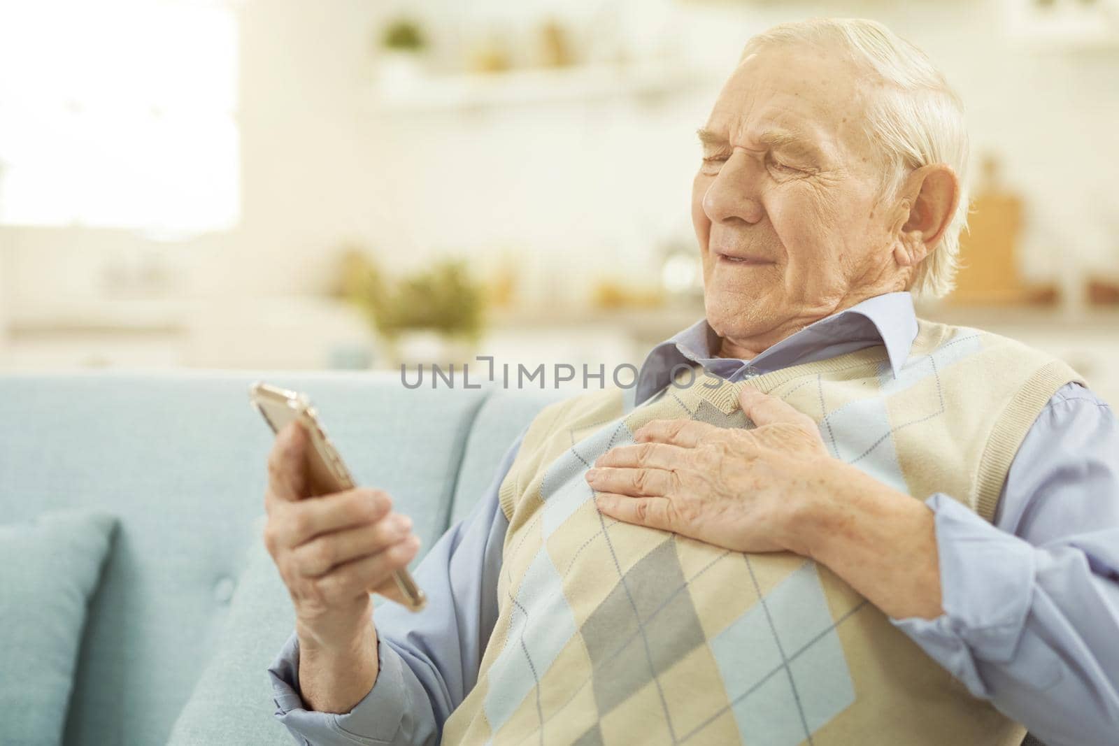 Sick Caucasian elderly man sitting on cozy sofa and using smartphone while talking online