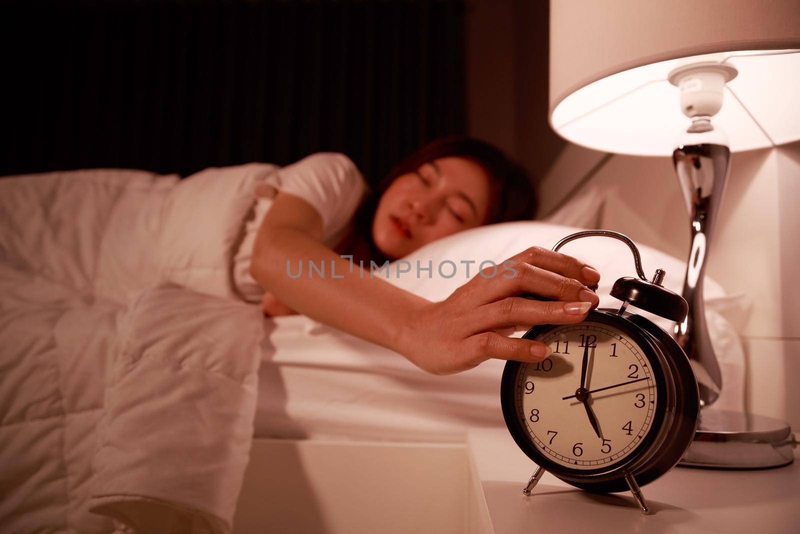 sleepy woman in bed with eyes closed extending hand to alarm clock in the morning by geargodz