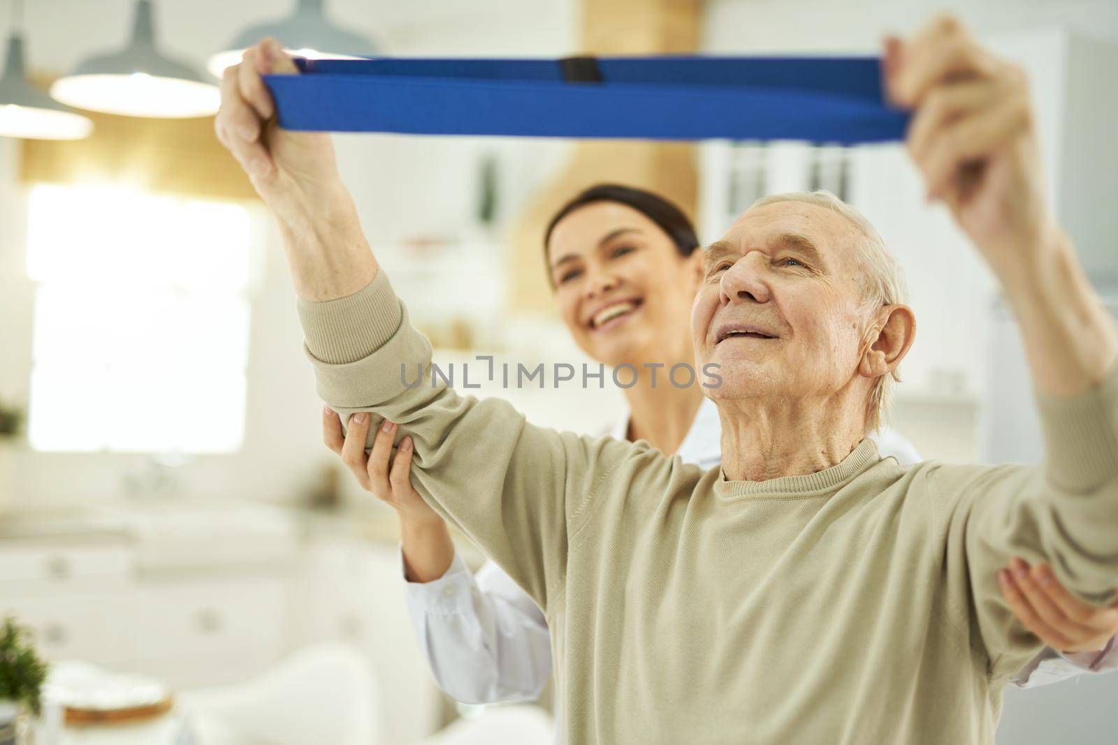 Senior citizen raising his hands up and doing exercises with fitness rubber band at home with doctor