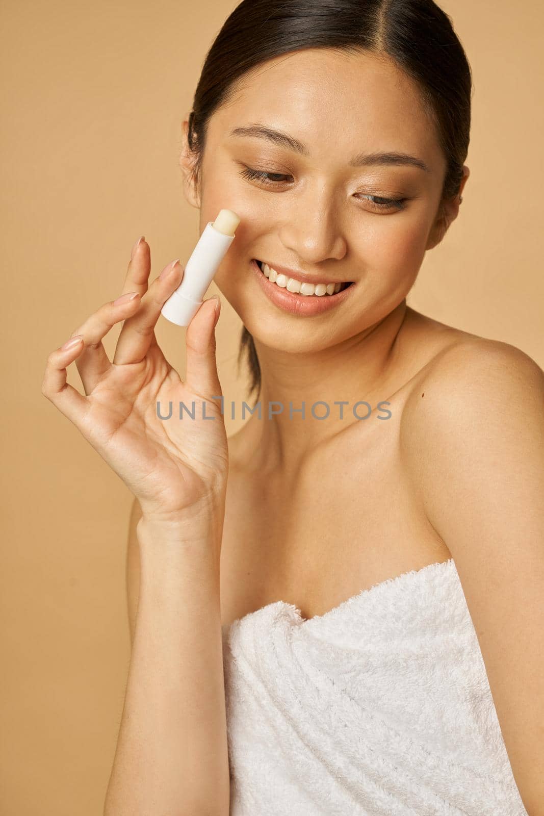 Attractive young woman wrapped in towel smiling and holding lip balm while posing isolated over beige background by friendsstock