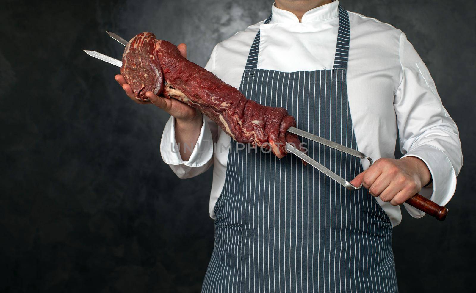 Crop cook in uniform and apron holding skewer with fresh raw meat on black background