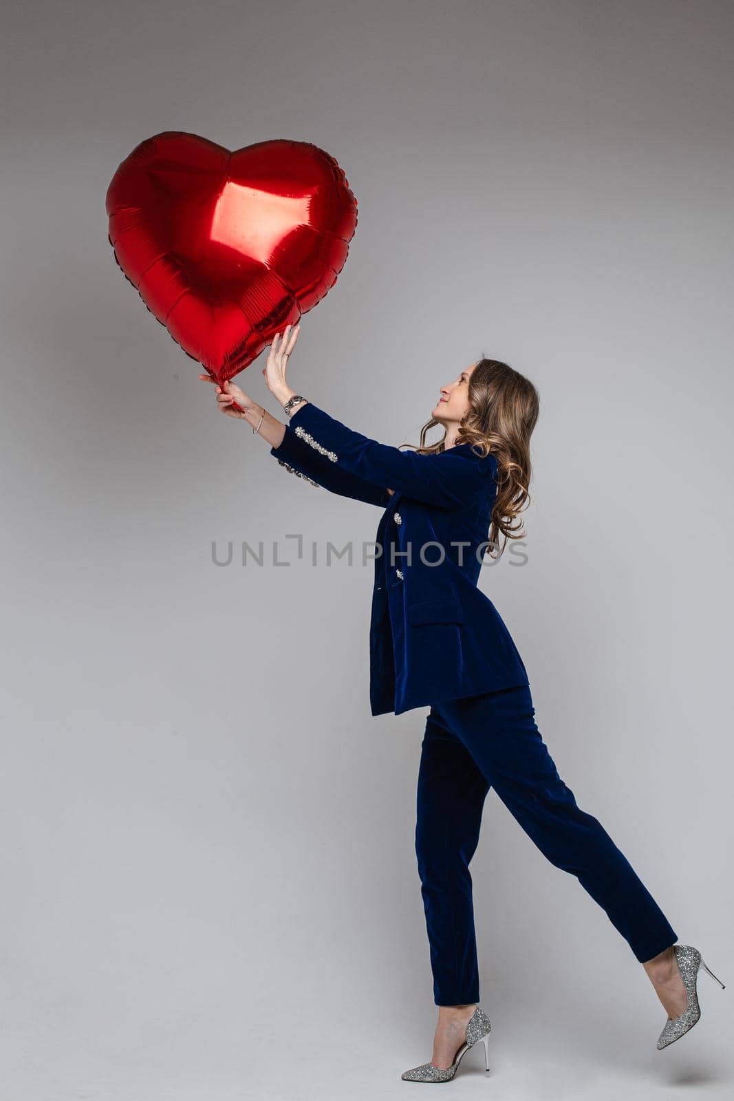 Full length of attractive Caucasian woman in blue velvet suit with jacket and trousers and high sparkling heels holding red heart-shaped symbol of love balloon. Isolated.