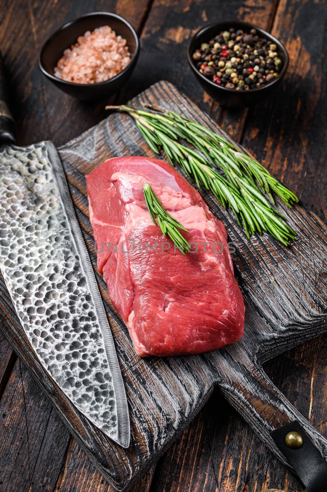 Raw lamb meat steak on a wooden cutting board with herbs. Dark wooden background. Top view.