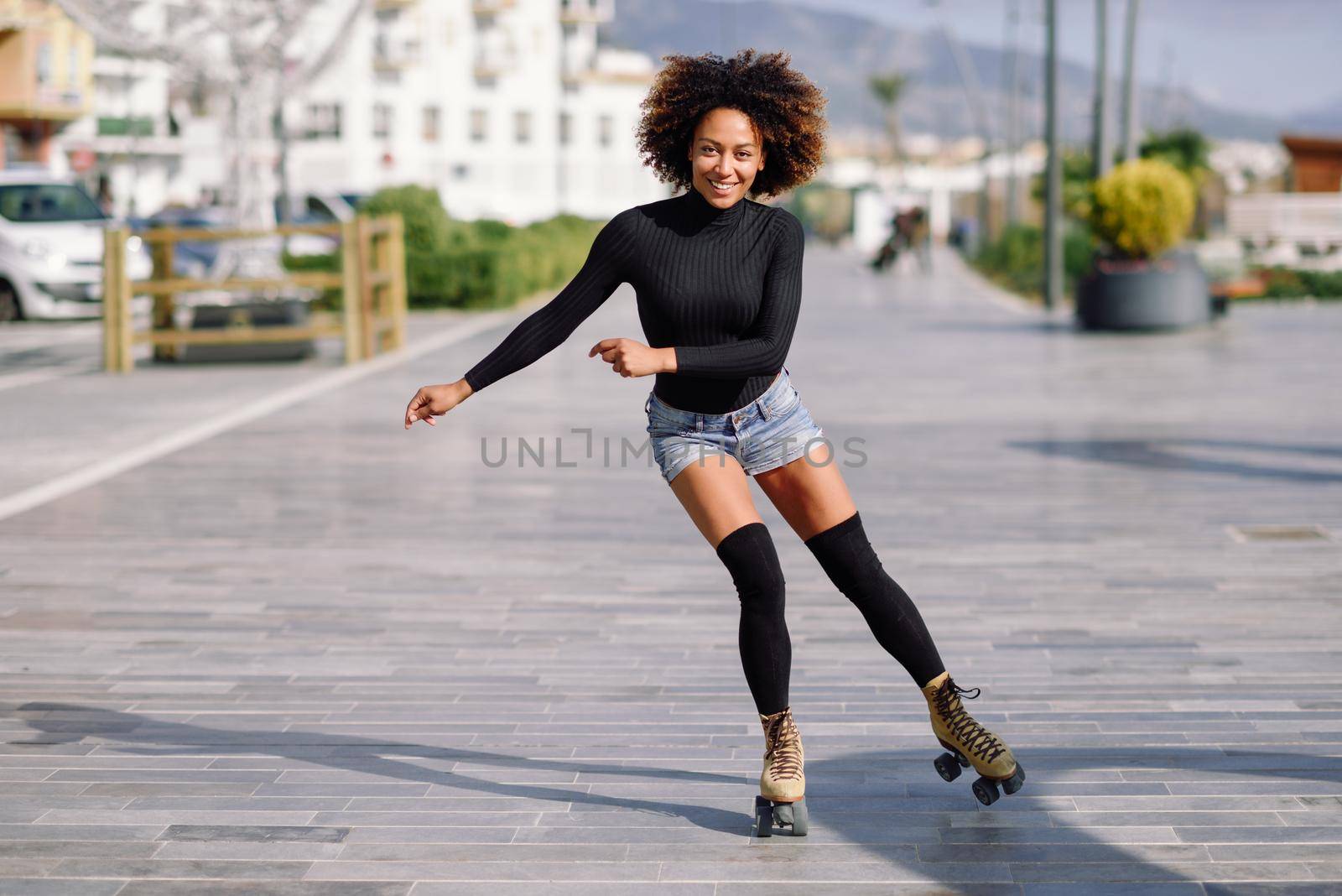 Young fit black woman on roller skates riding outdoors on urban street. Smiling girl with afro hairstyle rollerblading on sunny day