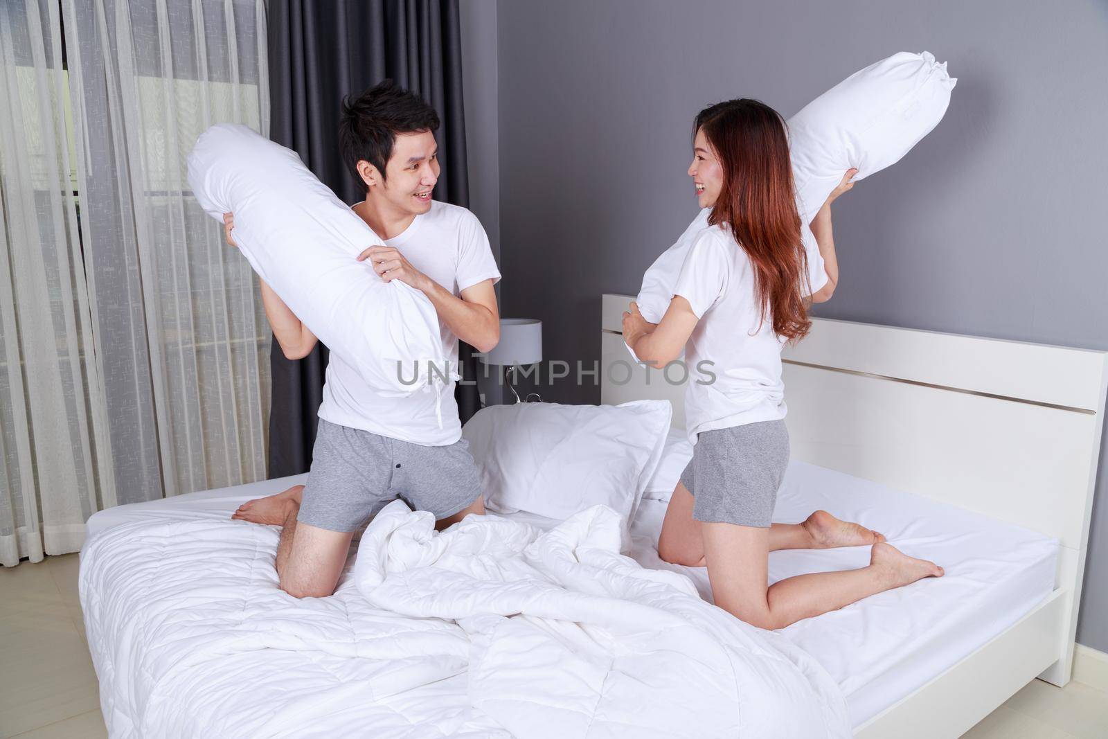 cheerful young couple having a bolster pillow fight on bed in bedroom by geargodz