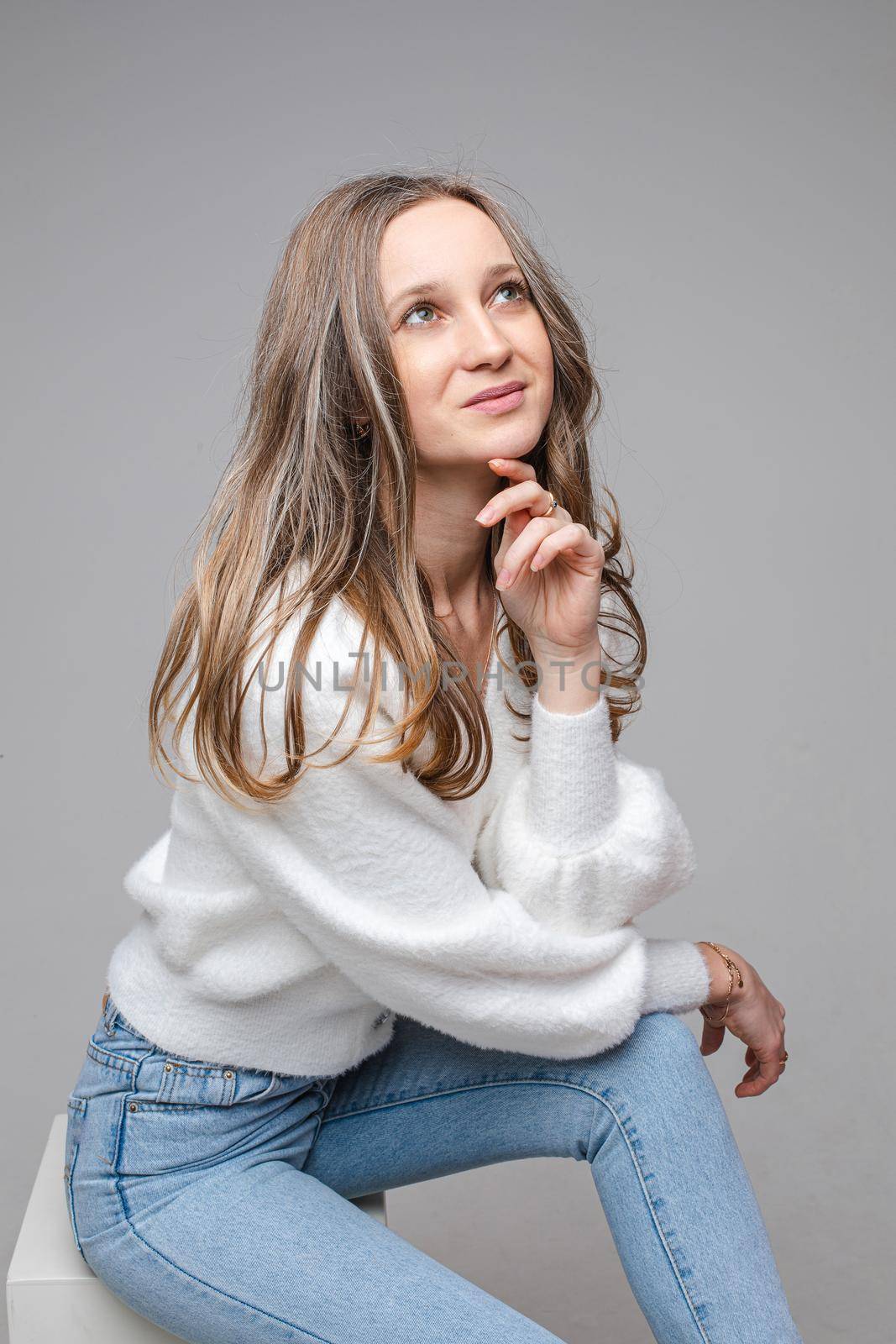 Portrait of young dreamy long-haired caucasian girl in white sweater and blue jeans on gray studio background by StudioLucky