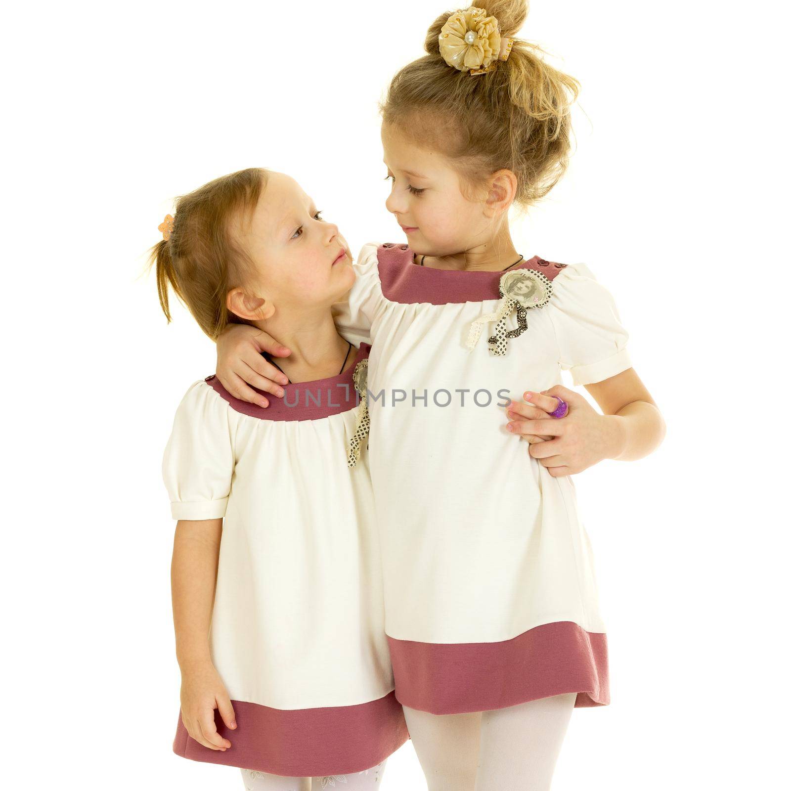 Cute adorable little sisters standing and hugging. Lovely girls dressed in the same dresses looking at each other against white background. Two happy kids embracing each other