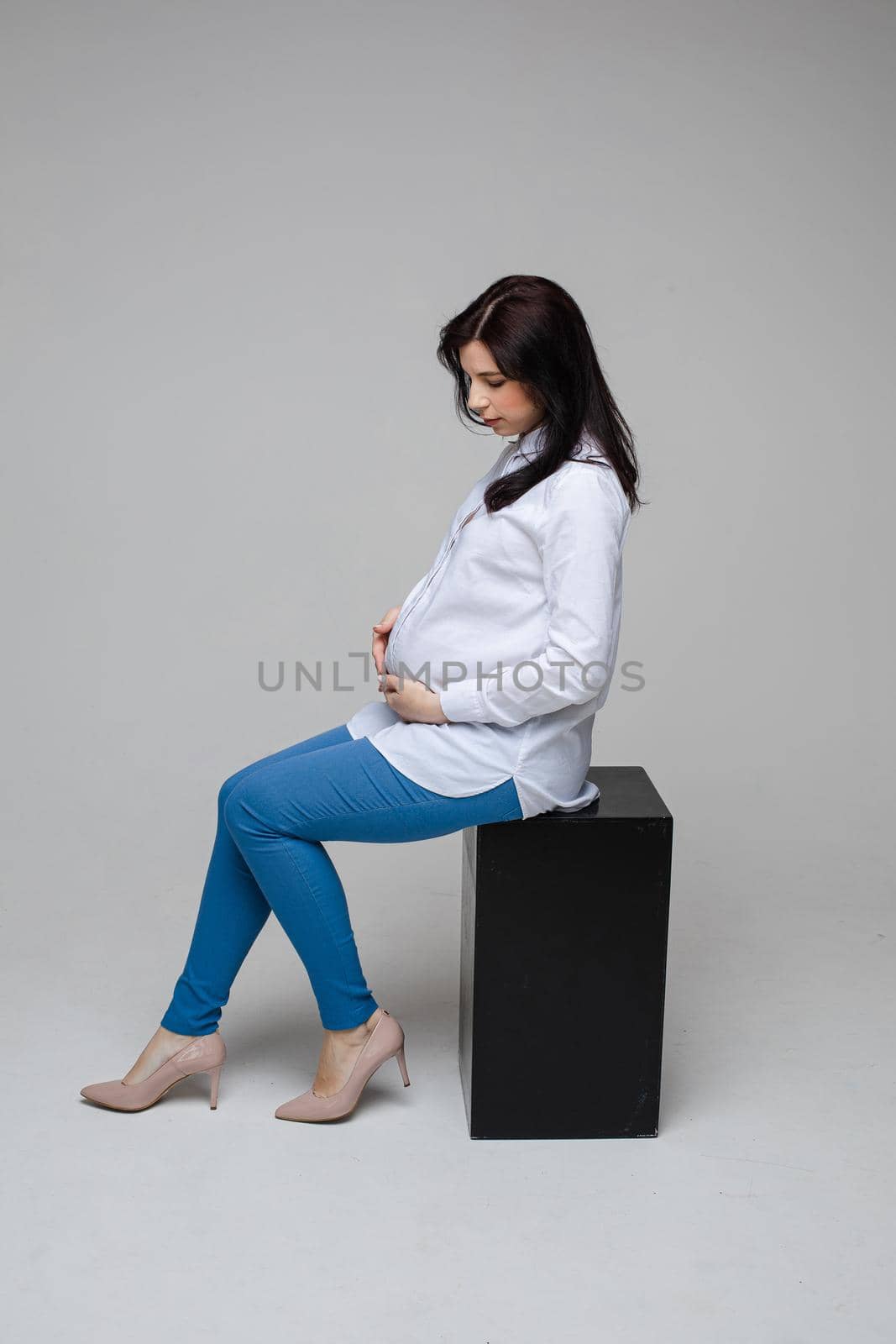 beautiful pregnant mum with black hair in a shirt and jeans sitting on a chair, picture isolated on grey background