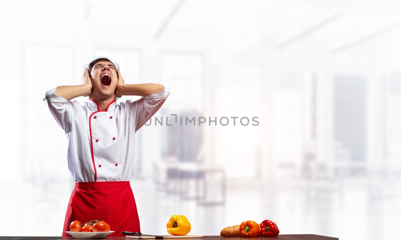 Young male chef screaming in shock and horror. Emotional caucasian chef in white hat and red apron standing with open mouth in light kitchen interior. Creative marketing campaign, big discounts.