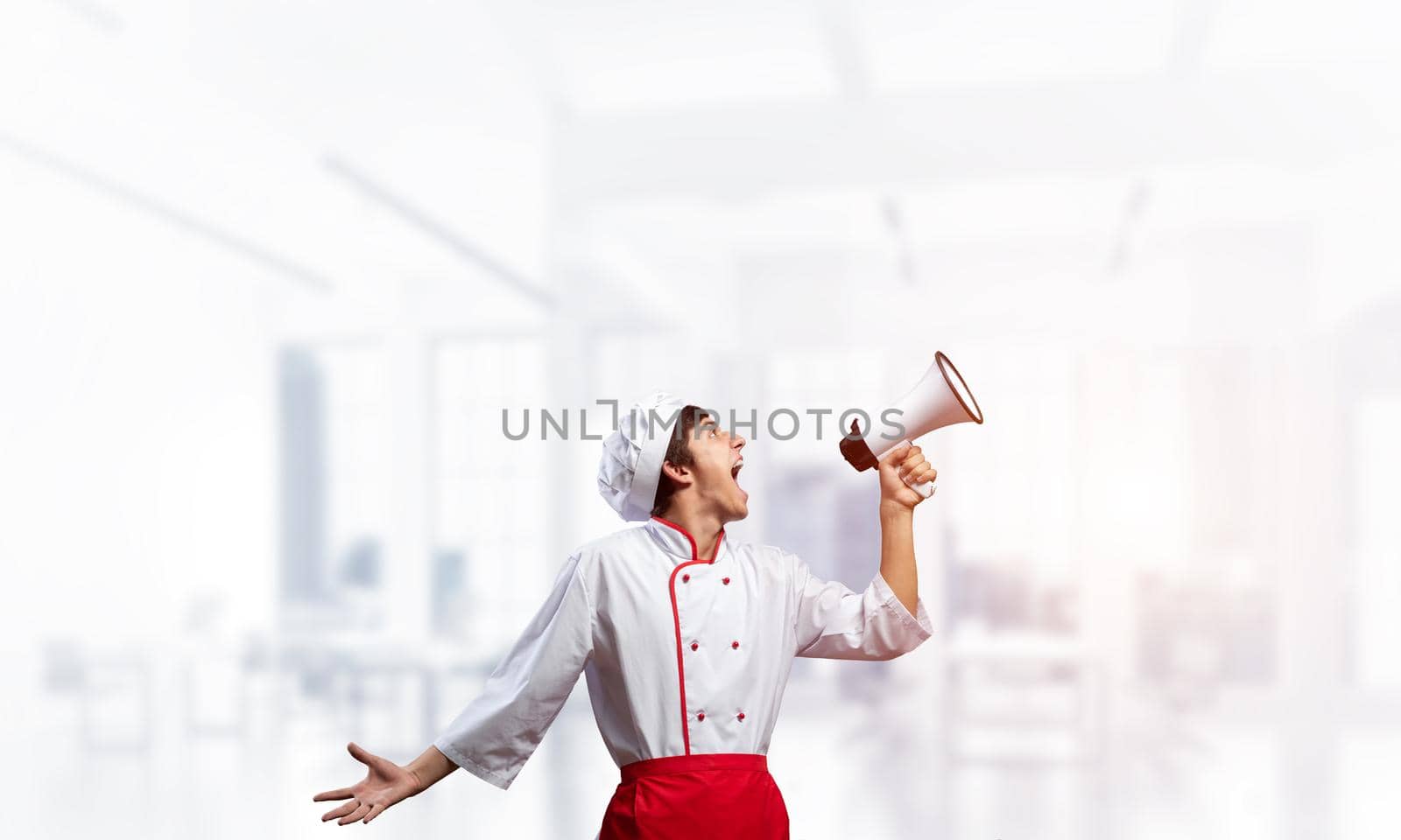 Young chef shouting loudly into megaphone. Emotional caucasian chef in white hat and red apron on light blurred background. Restaurant advertisement, food retail campaign announcement.