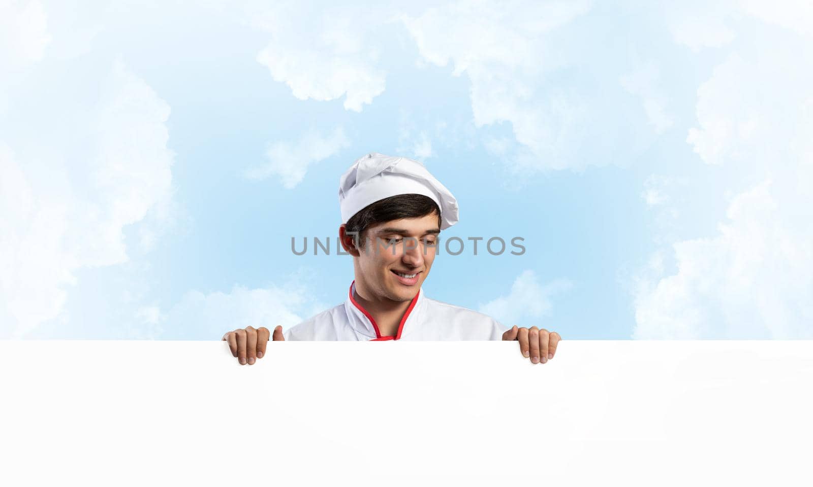 Young male chef with blank signboard. Handsome chef in white and red uniform on blue sky background. Cooking classes announcement. Professional catering service concept with copy space.