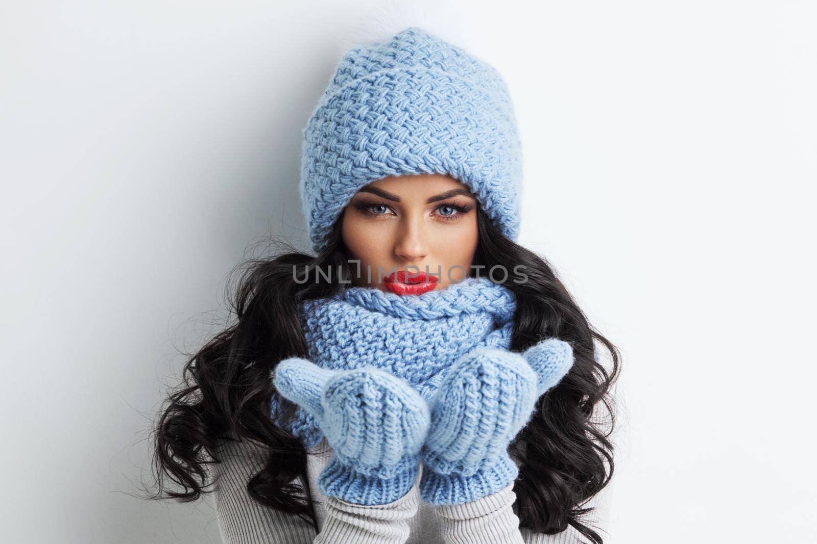 Beautiful woman in blue winter hat, scarf and mittens blowing on palms, white background