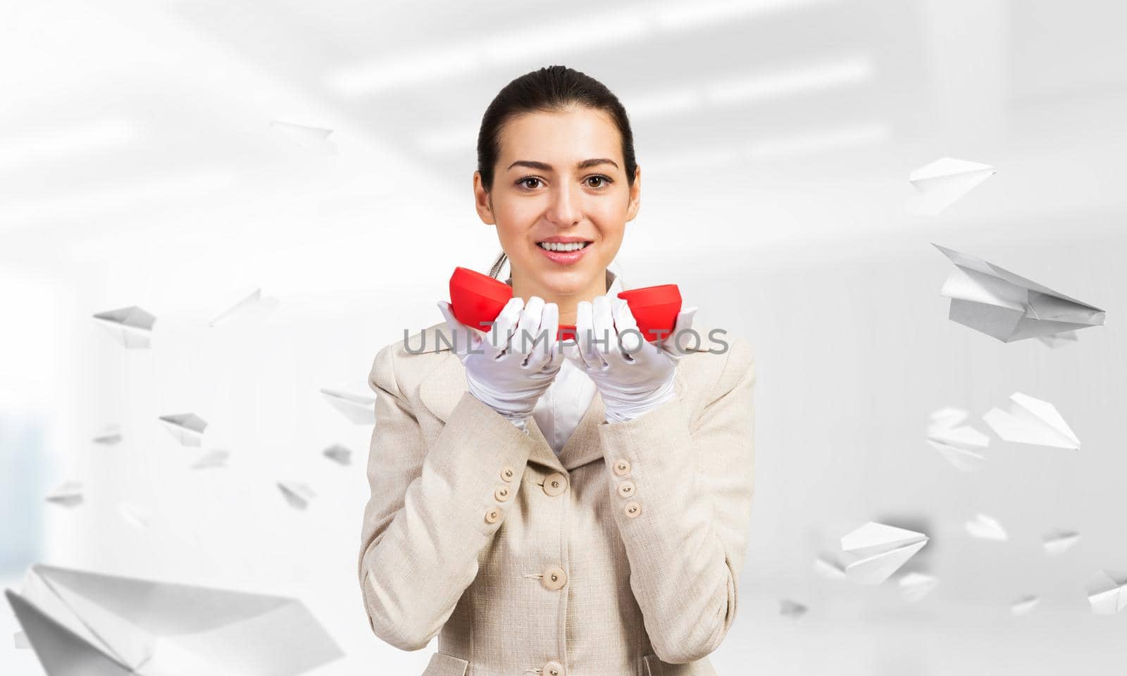 Smiling young woman holding retro red phone in office with flying paper planes. Call center operator in business suit with landline phone. Hotline telemarketing. Business assistance and consultation.