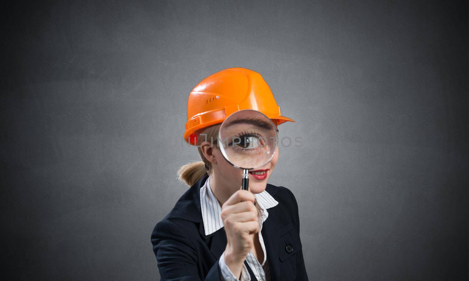 Technical inspector looking through magnifying glass. Woman civil engineer in orange safety helmet checking with magnifier on grey wall background. Commercial real estate appraisal and consultation.
