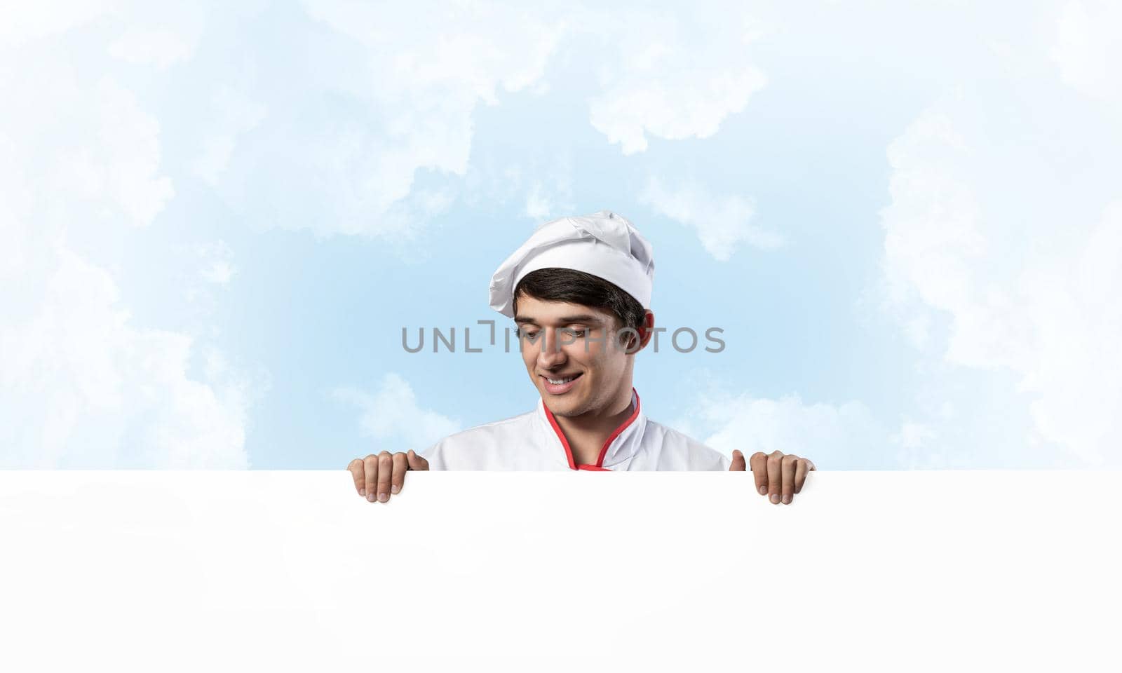 Young male chef with blank signboard. Handsome chef in white and red uniform on blue sky background. Cooking classes announcement. Professional catering service concept with copy space.