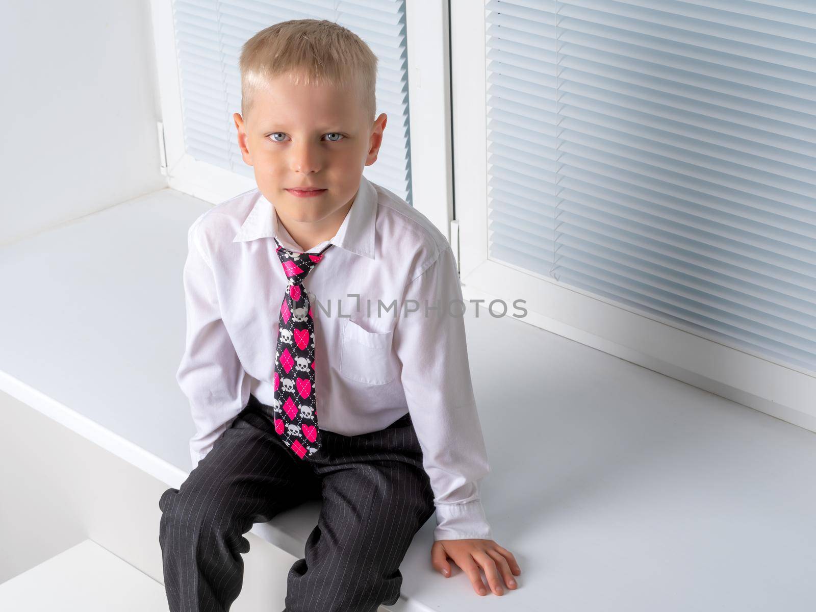 A cute little boy sits near a window closed by horizontal white blinds on the windowsill. It is possible to use the concept for advertising plastic windows and blinds.