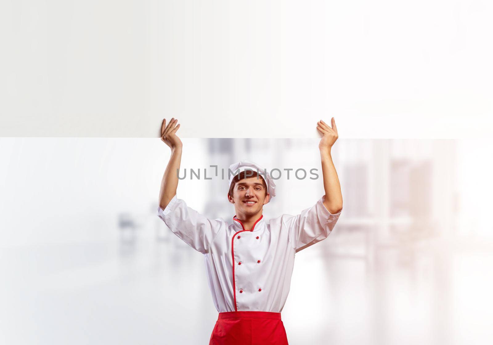 Young male chef with blank signboard above head. Chef in white and red uniform on light room blurred background. Cooking classes announcement. Professional catering service concept with copy space