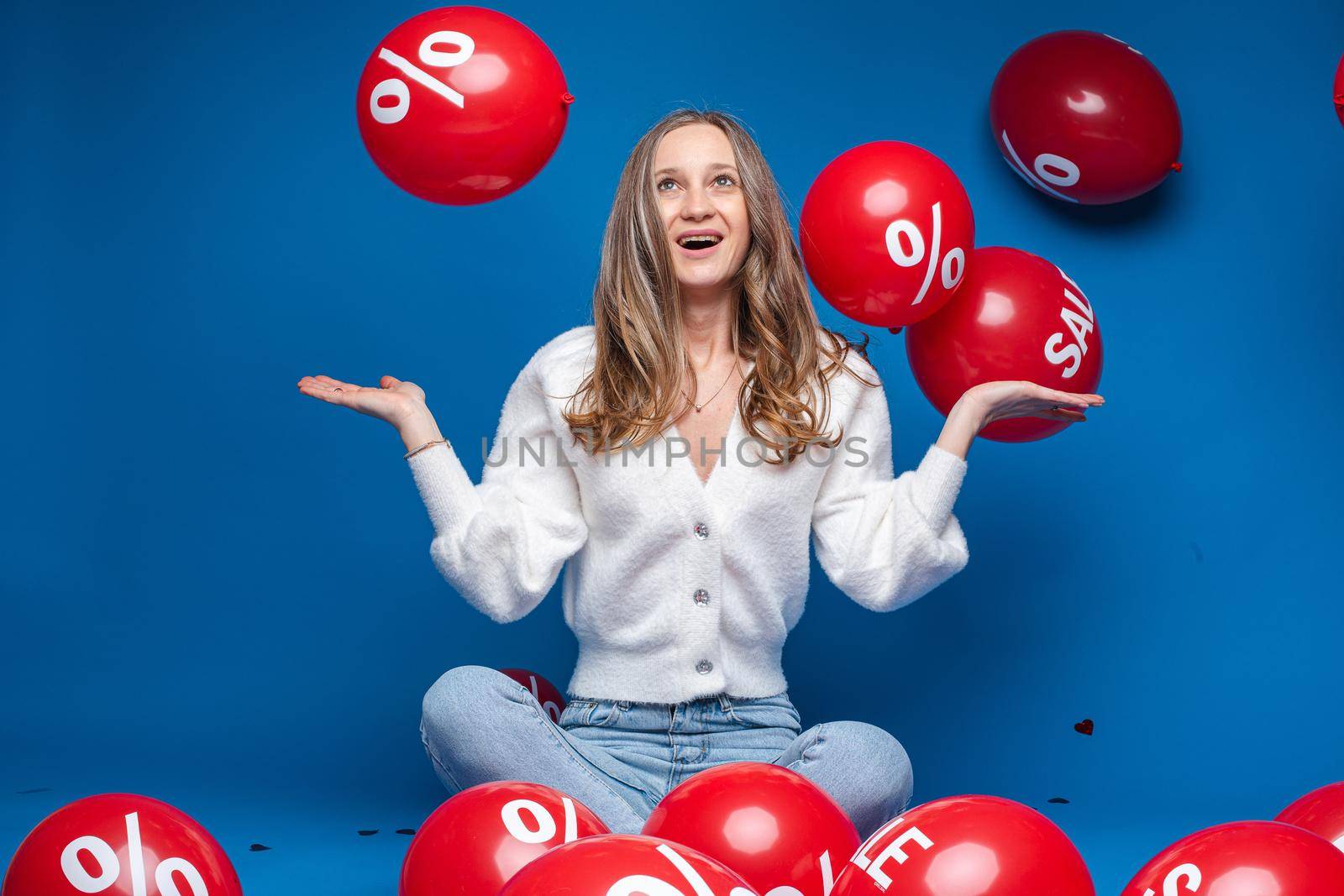 Studio portrait of happy fair-haired girl in white cardigan and jeans holding arms half bent and looking at flying red balloons with sale word and percent sign. Shopping spree, sales concept.