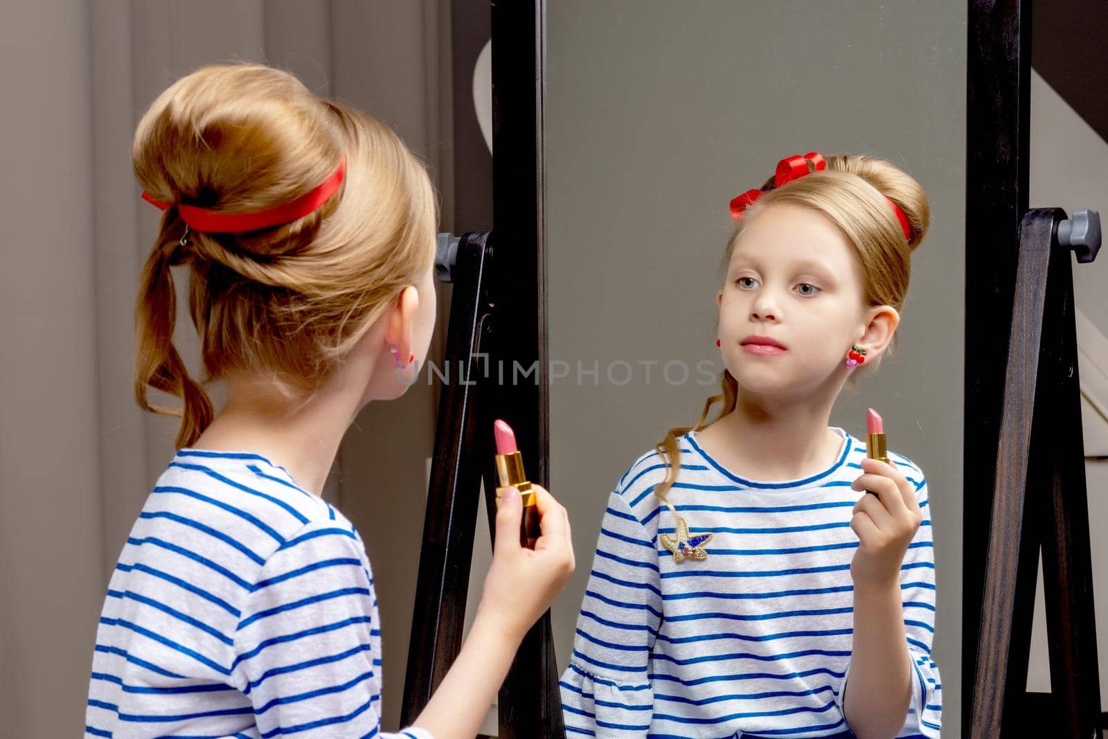 A nice little girl near the big mirror paints her lips with lipstick. The concept of childhood, style and fashion.