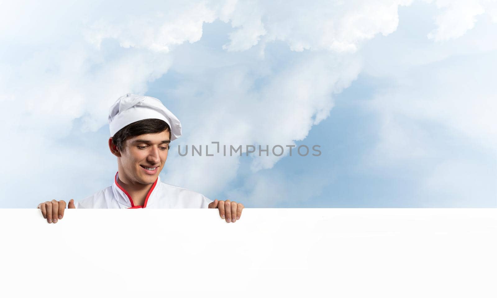 Young male chef with blank signboard. Handsome chef in white and red uniform on blue sky background. Cooking classes announcement. Professional catering service concept with copy space.