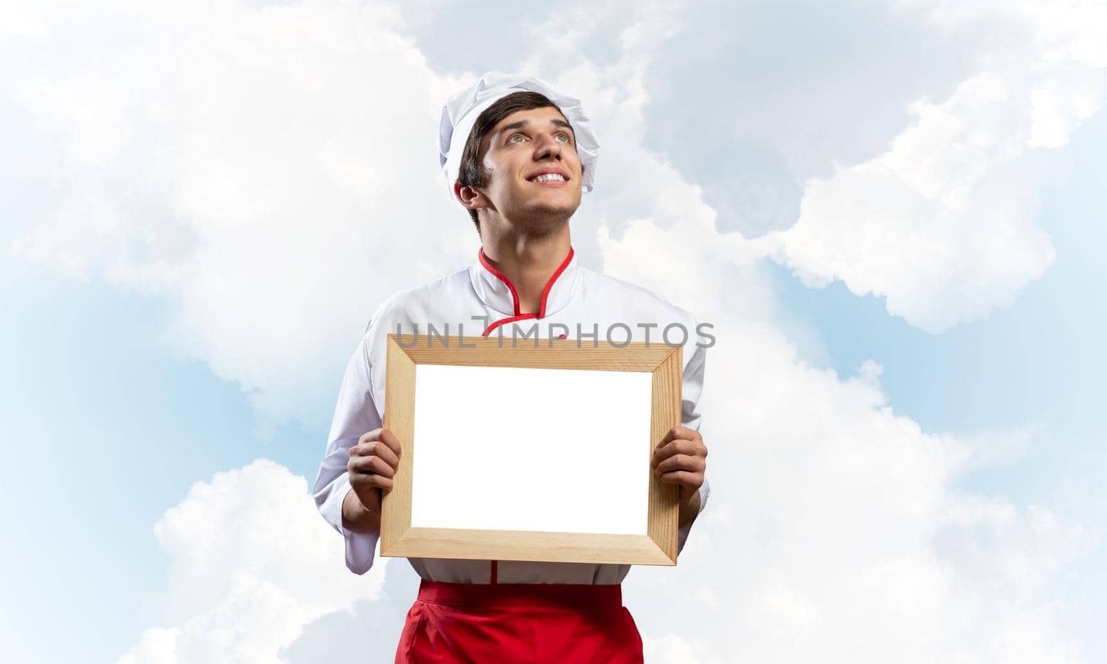 Young male chef holding blank white board for text. Handsome chef in white and red uniform on blue sky background. Cooking classes advertising. Professional catering service concept with copy space.