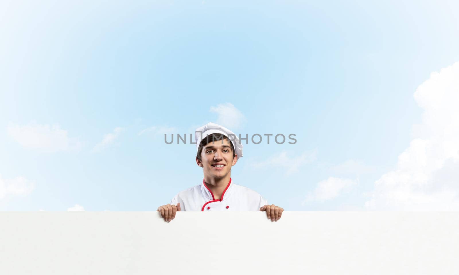 Young male chef with blank signboard. Handsome chef in white and red uniform on blue sky background. Cooking classes announcement. Professional catering service concept with copy space.