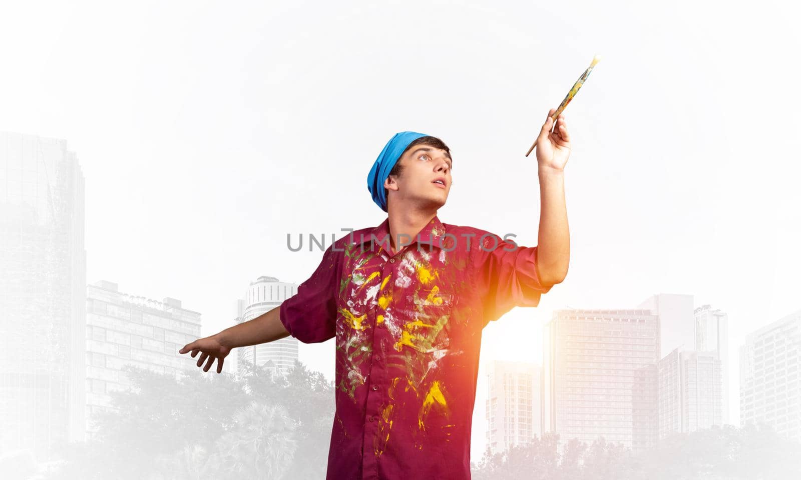 Young emotional artist gesturing with paintbrush. Happy painter in shirt and bandana standing on background modern office buildings. Creative hobby and artistic occupation concept with copy space