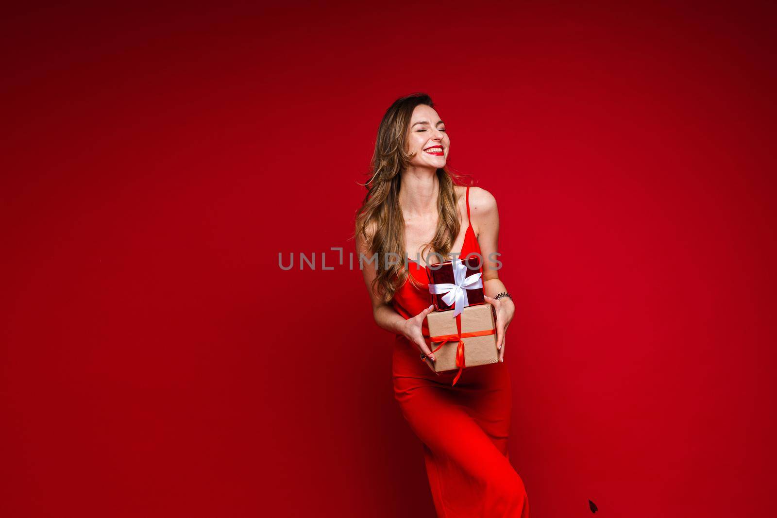 Happy young woman having fun while holding gift boxes, isolated on red background. Saint Valentine Day concept