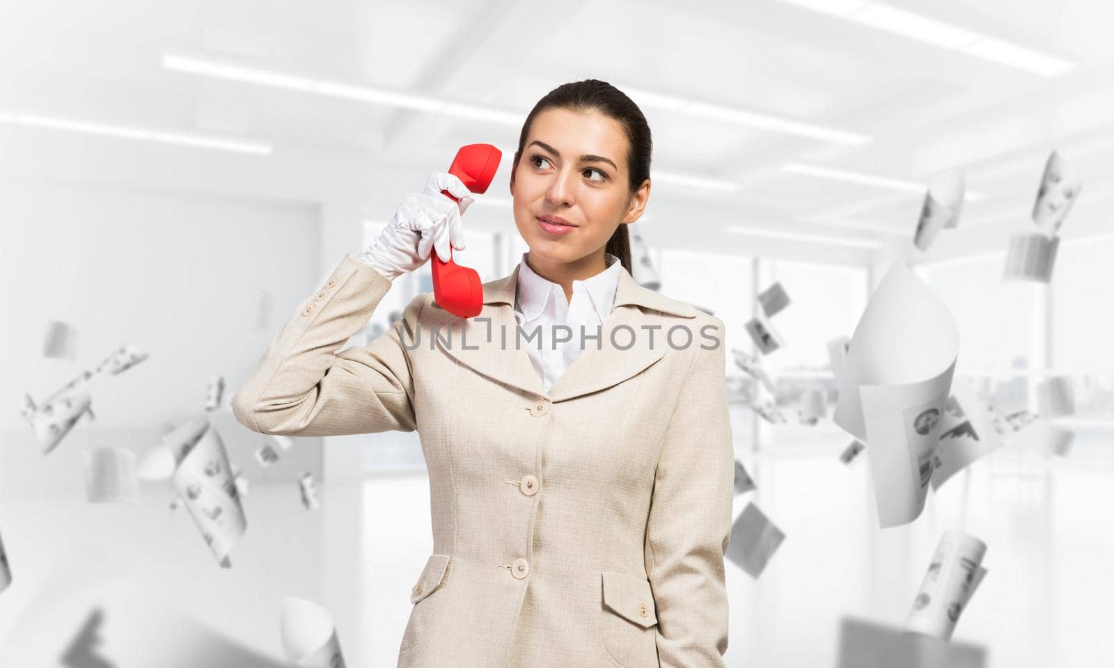 Smiling young woman holding retro red phone in office with flying paper documents. Call center operator in business suit with telephone. Hotline telemarketing. Business assistance and consultation.