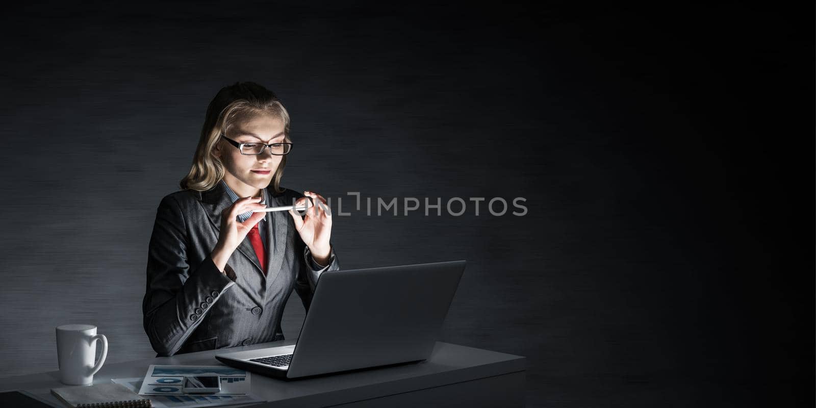Attractive young businesswoman in glasses sitting at office desk and working at laptop. Beautiful lady in business suit and tie busy at work in evening. Career building and business development