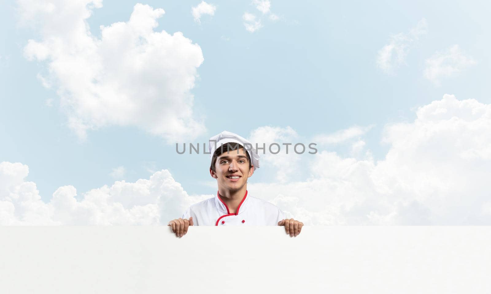 Young male chef with blank signboard. Handsome chef in white and red uniform on blue sky background. Cooking classes announcement. Professional catering service concept with copy space.