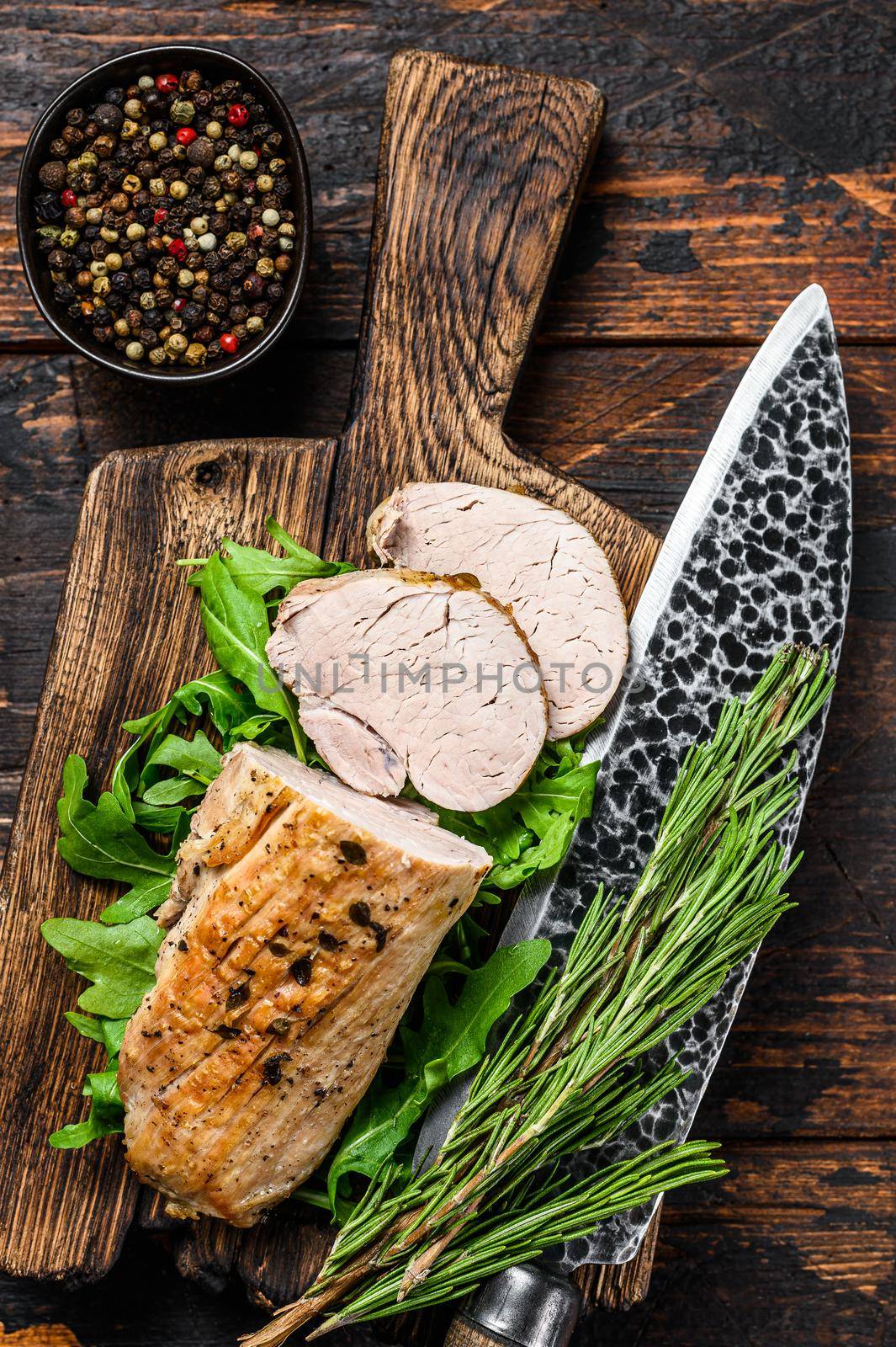 Cut baked pork tenderloin steak on a cutting board with arugula. Dark background. Top view.