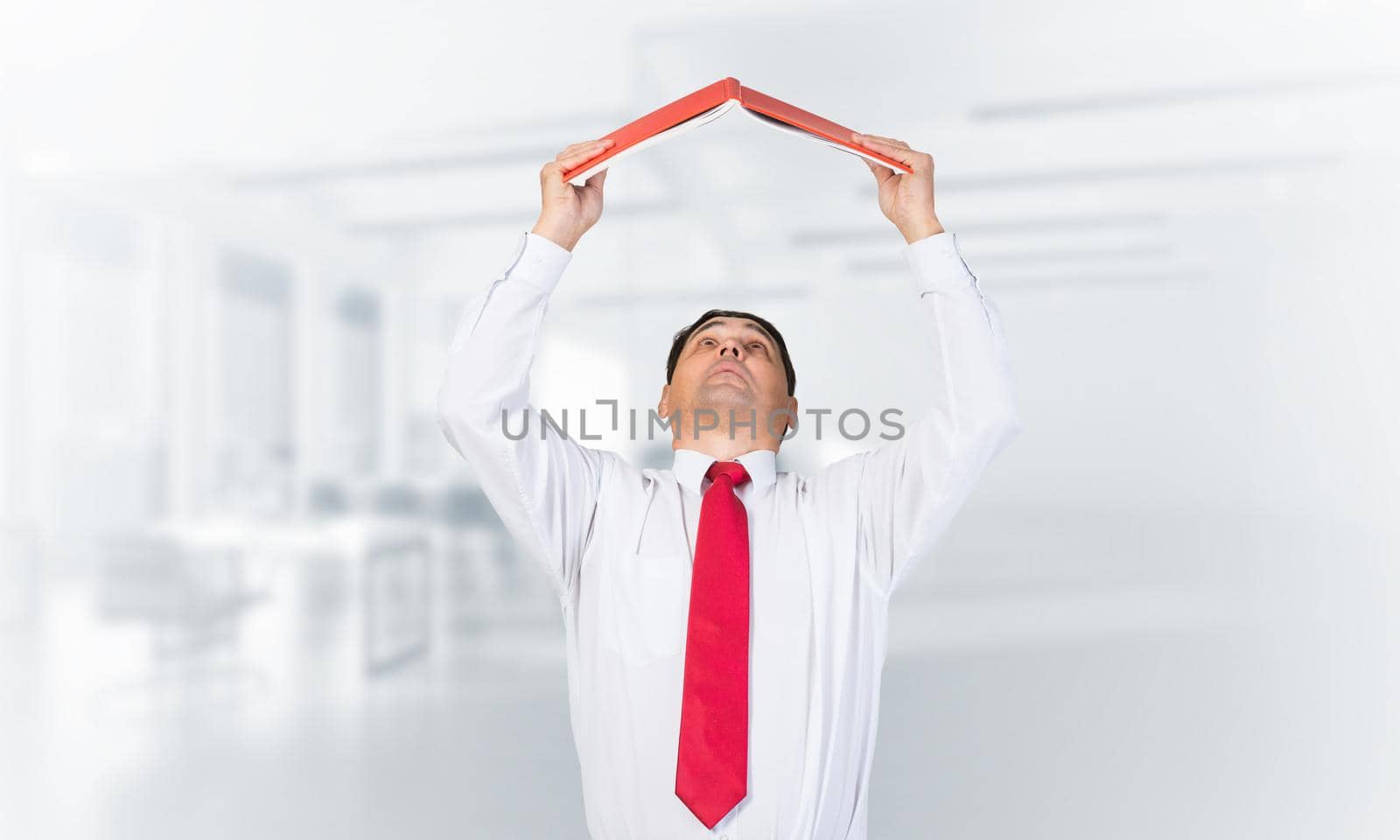 Senior businessman holding open book overhead as roof. Portrait of adult man in white shirt and tie standing in blurred office interior. Education and professional knowledges. Reference information.