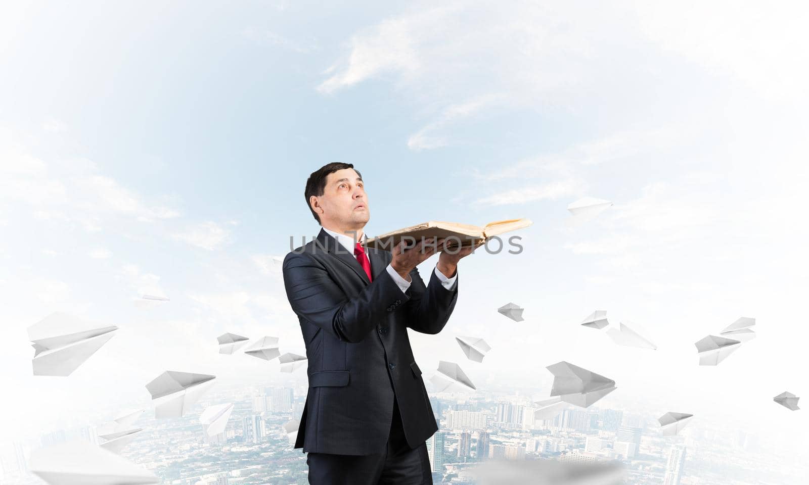 Serious businessman with open book looking up. Man in suit standing on downtown background with flying paper planes. Lawyer holding big legal regulation book. Business accounting and consulting.