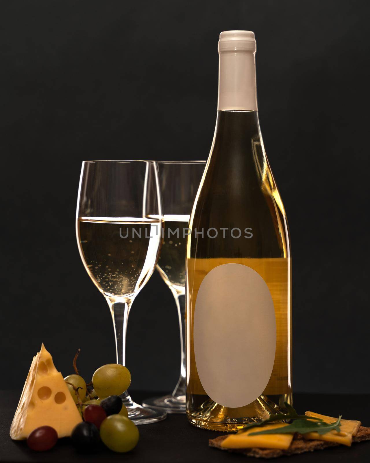 On a black background, a bottle of white wine, two glasses of white wine, next to grapes, a piece of triangular cheese, a sandwich