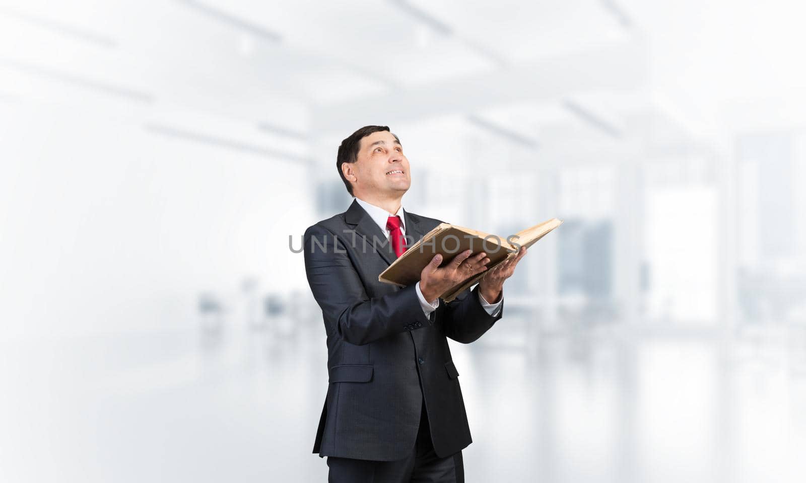 Smiling businessman with open book looking up. Adult man in business suit standing in blurred office interior. Lawyer holding big legal regulation book. Professional business accounting and consulting