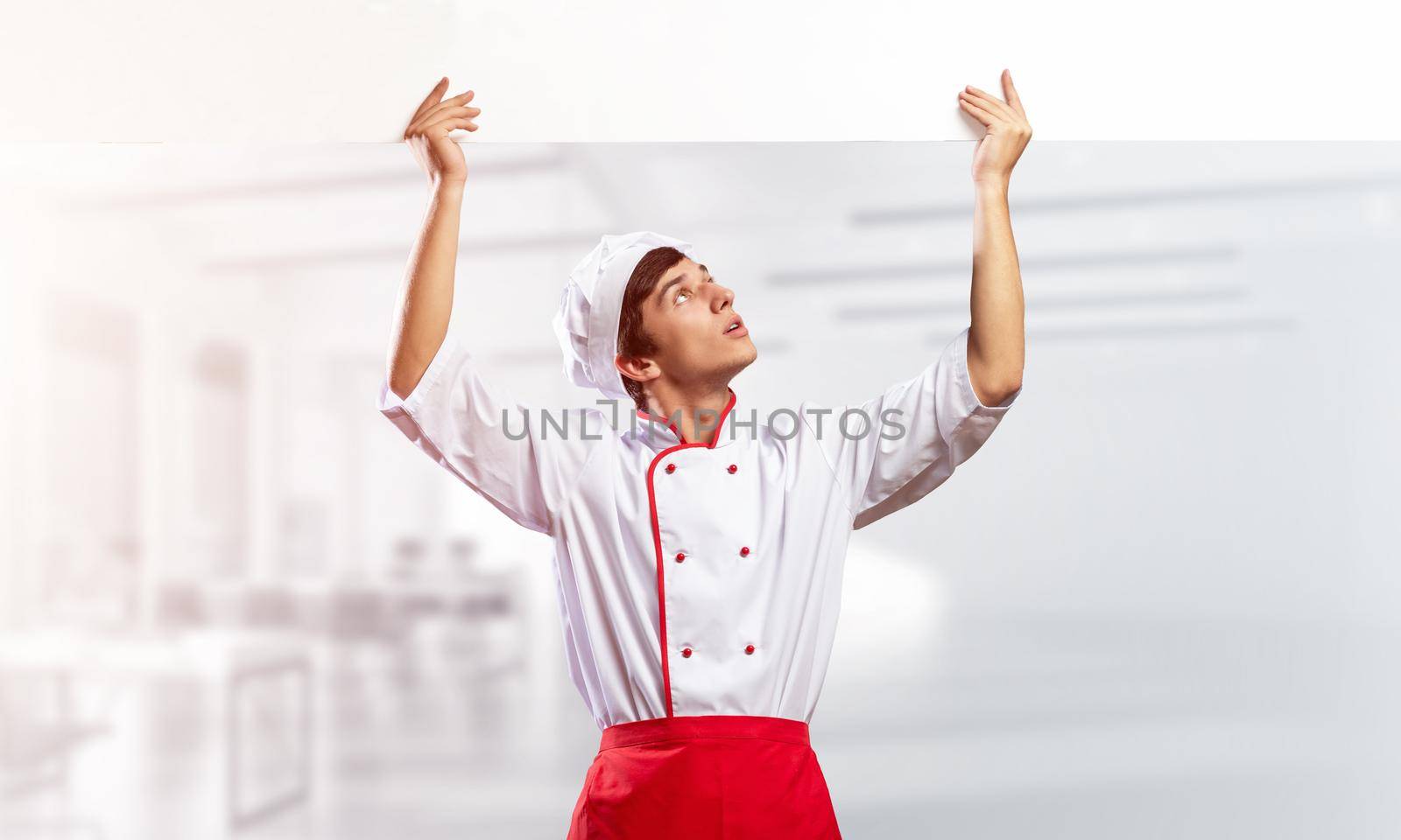 Young male chef with blank signboard above head. Chef in white and red uniform on light room blurred background. Cooking classes announcement. Professional catering service concept with copy space