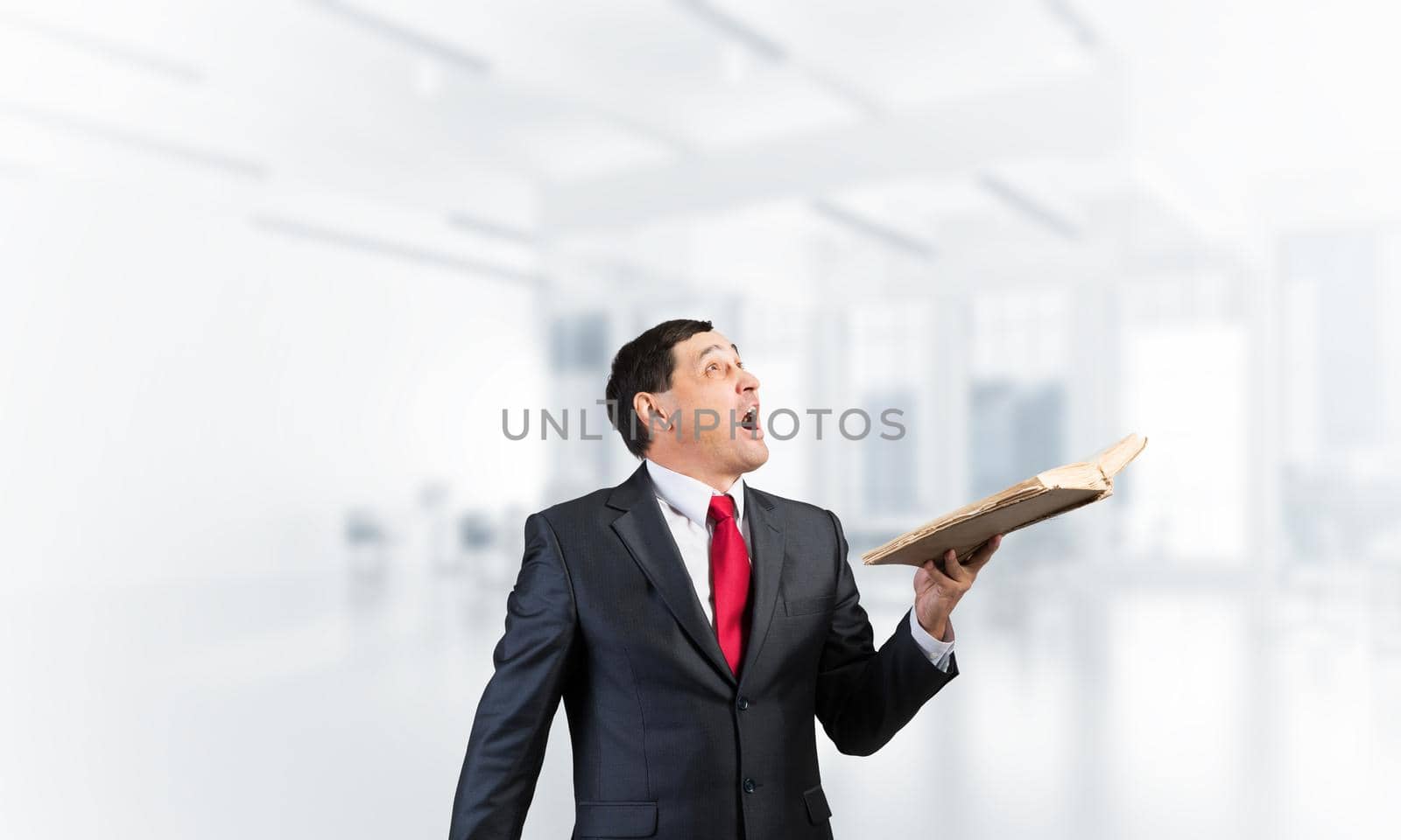 Surprised businessman holding open book and looking up. Startled adult man in business suit and tie standing in blurred office interior. Education and knowledges. New and actual information concept