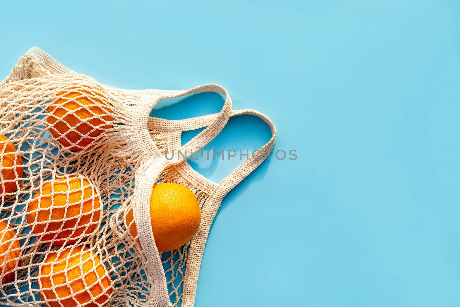 Mesh white cotton reusable bag with oranges on a blue background. Environmentally friendly shopping, the environment and the concept of healthy foods, citrus fruits with vitamin C. Place for text.