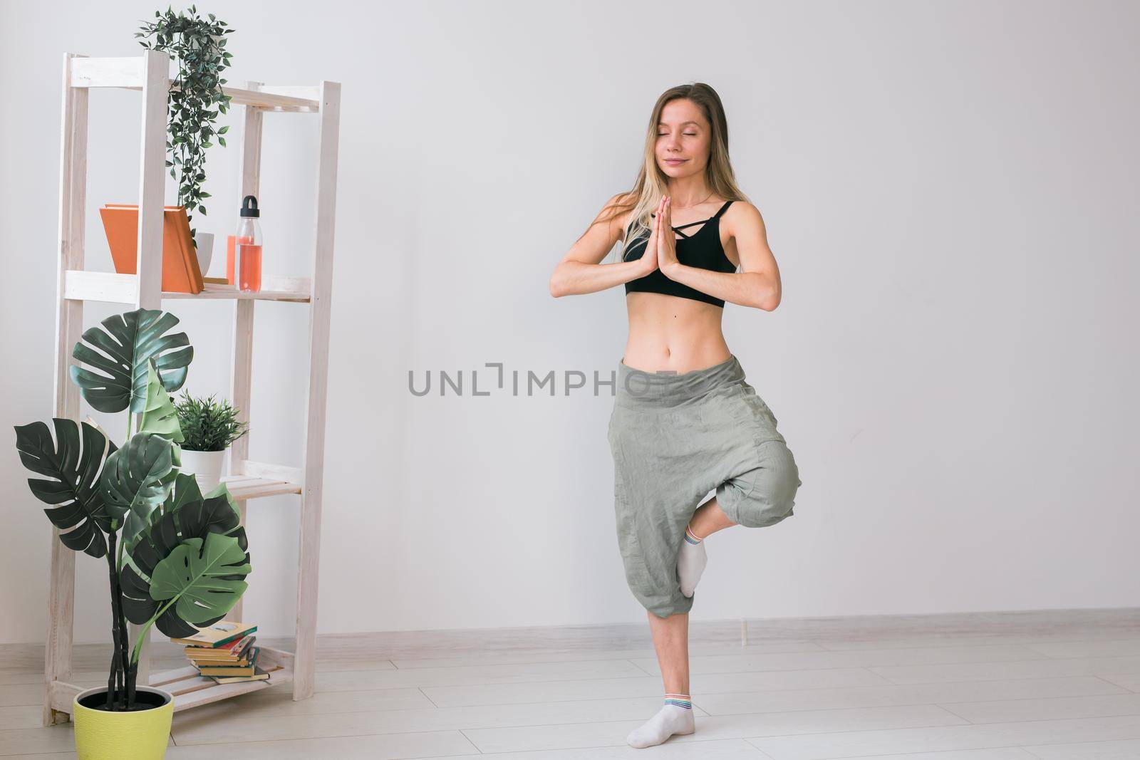 Woman practise yoga performs. Tree pose standing on mat inside of cozy room with plants and greenery. Healthy lifestyle