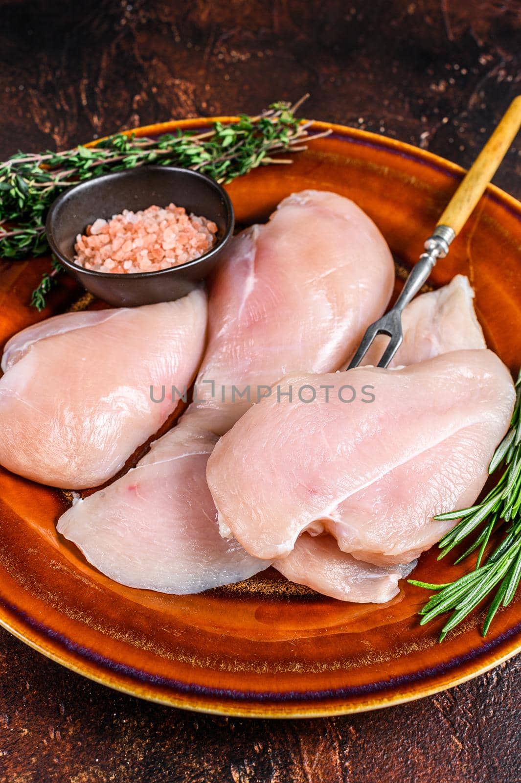 Raw slices of chicken breast fillet steaks on a rustic plate with herbs. Dark background. Top view by Composter