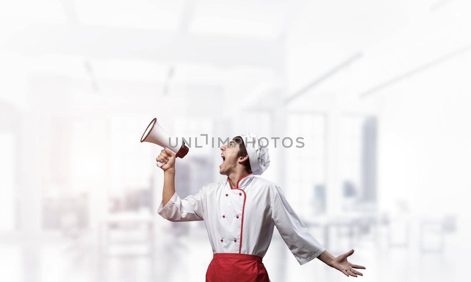 Young chef shouting loudly into megaphone. Emotional caucasian chef in white hat and red apron on light blurred background. Restaurant advertisement, food retail campaign announcement.