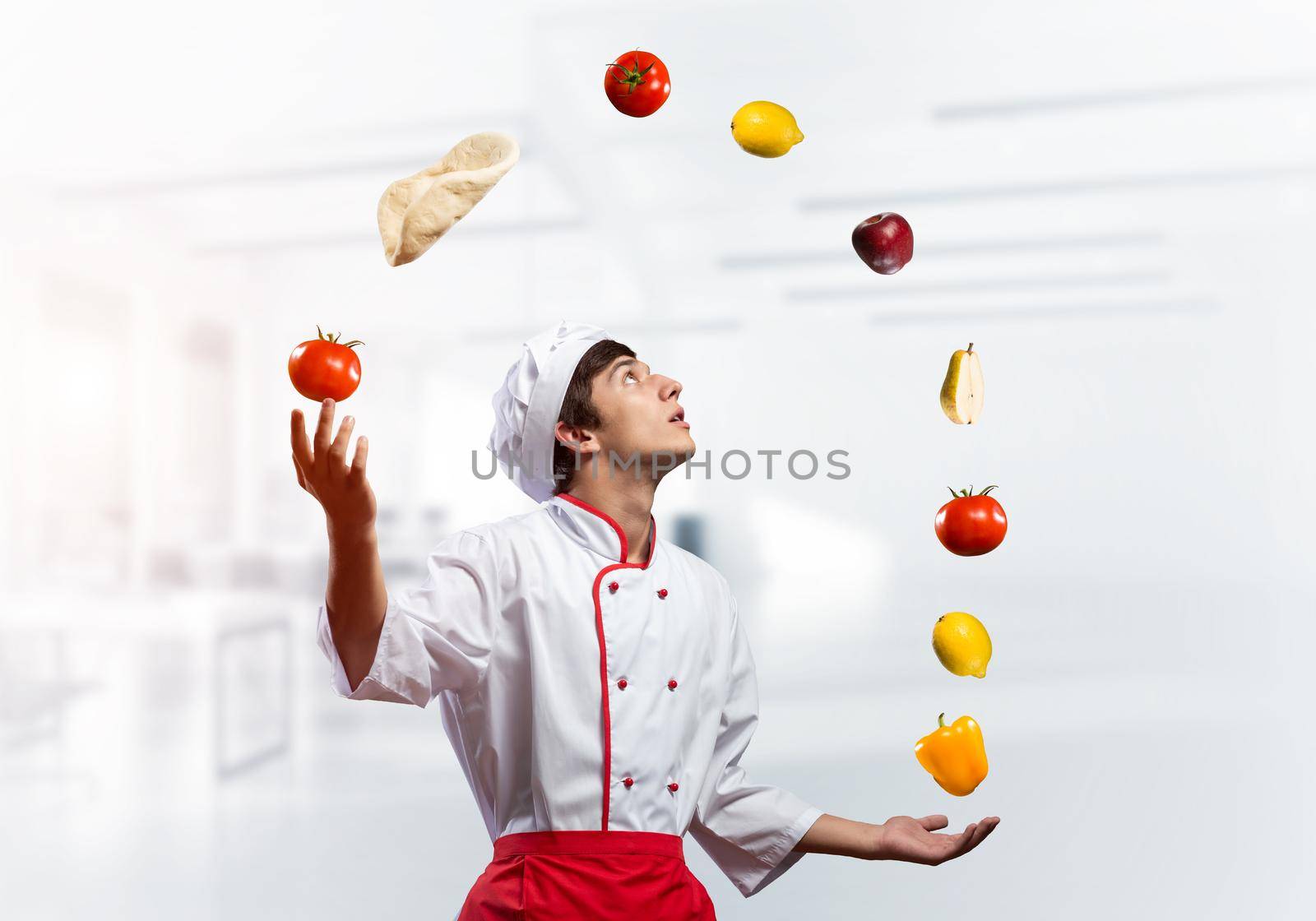 Young male chef juggles with food ingredients. Handsome chef in white hat and red apron in light kitchen interior. Professional cooking classes advertising. Food retail campaign announcement.