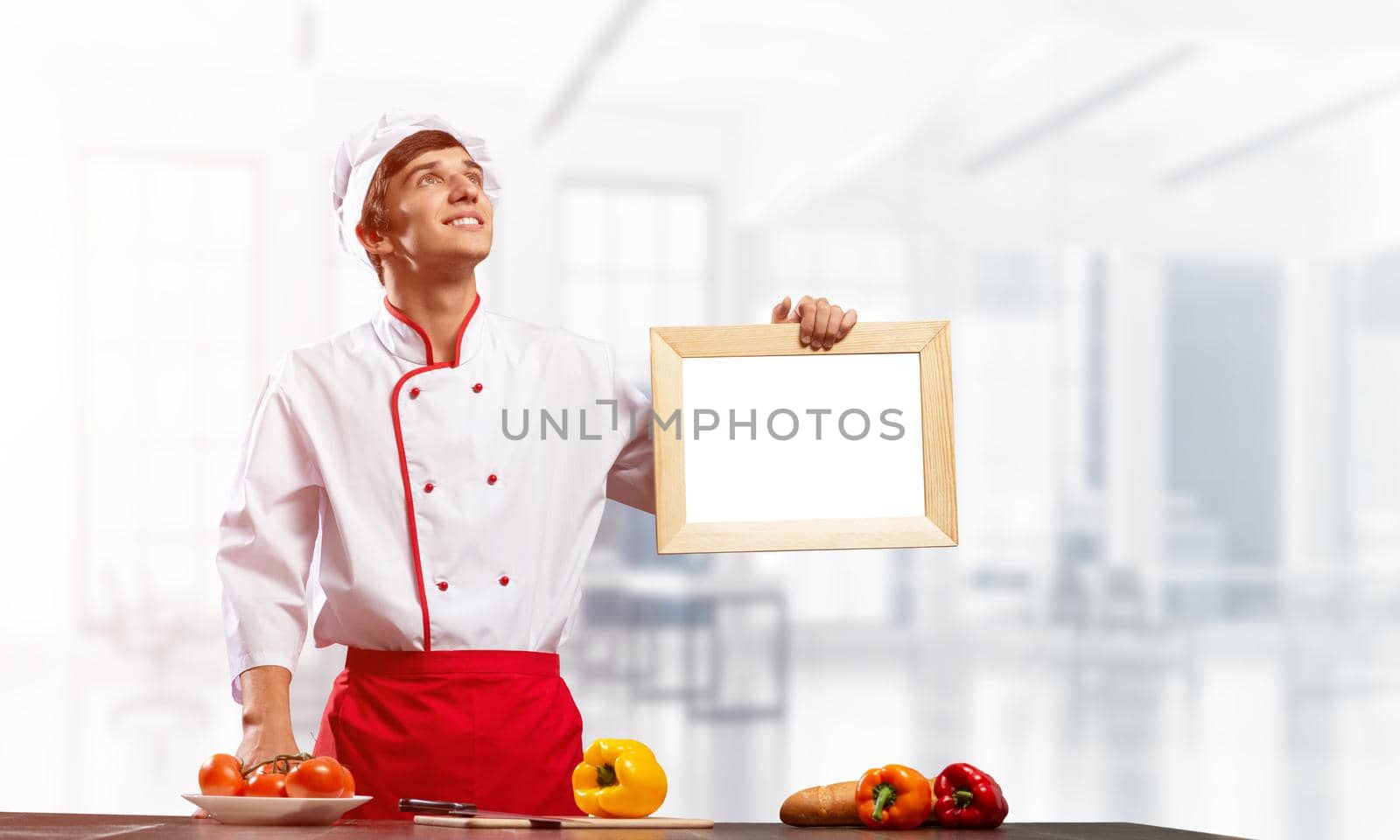 Young male chef standing near cooking table and holding blank white board. Handsome chef in white and red uniform in light kitchen interior. Cooking classes advertising. Professional catering service