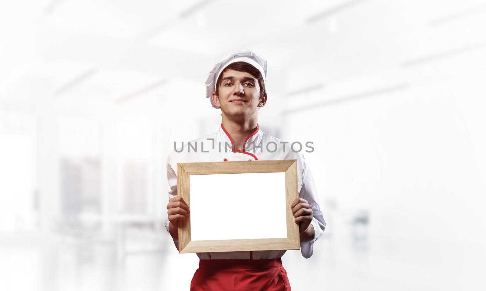 Young male chef standing near cooking table and holding blank white board. Handsome chef in white and red uniform in light kitchen interior. Cooking classes advertising. Professional catering service