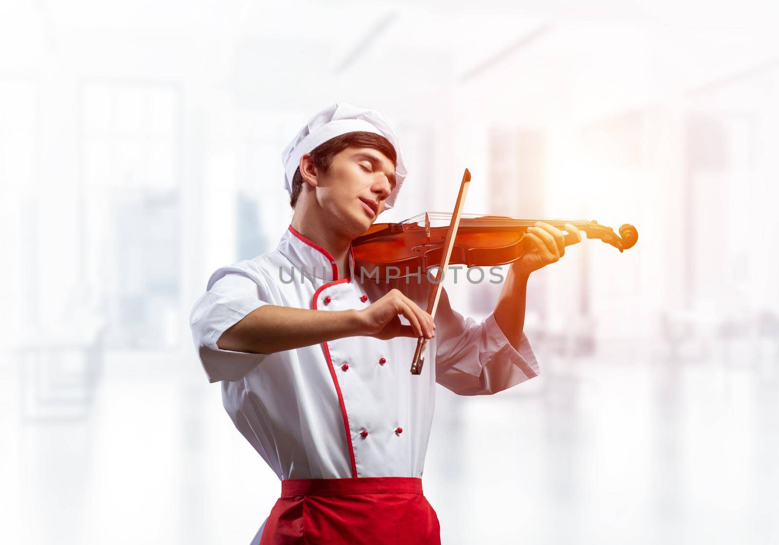 Young male chef with violin standing near cooking table with vegetables. Handsome chef in white hat and red apron in light kitchen interior. Creative culinary performance. High cooking skills concept