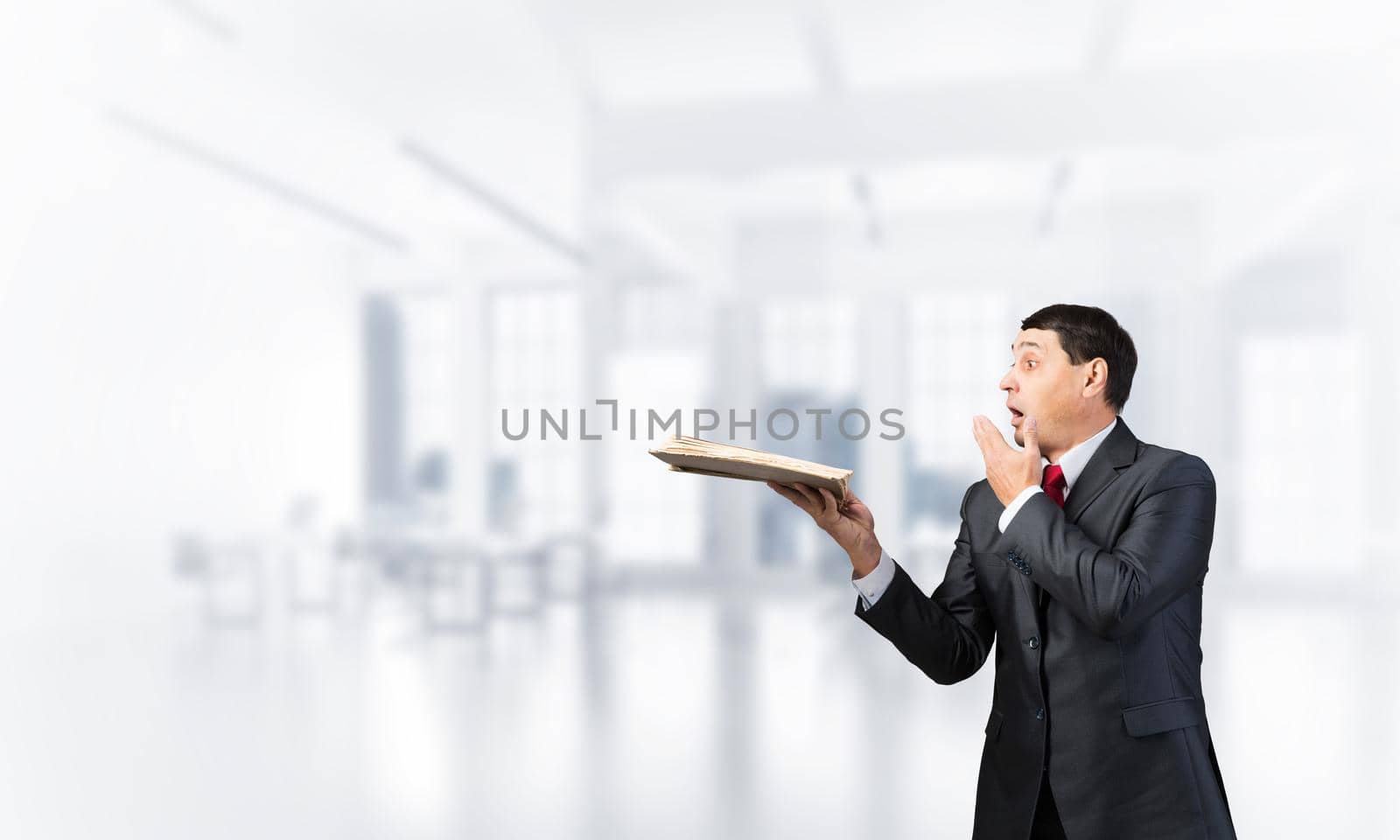Scared businessman covering mouth with hand and holding open book. Startled adult man in business suit and tie standing on blurred office background. Education and knowledges. Business accounting
