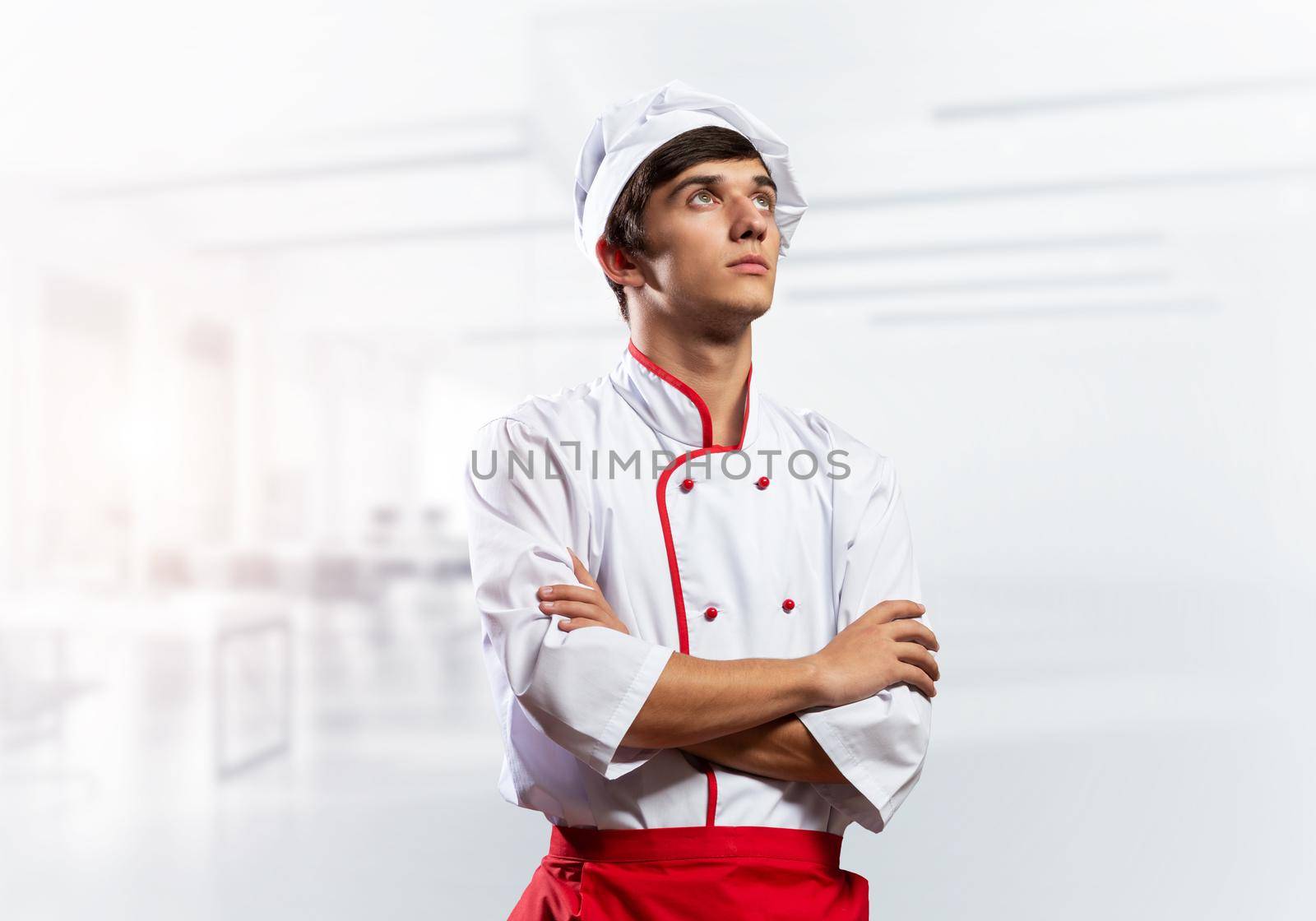 Young male chef standing with folded arms near cooking table with vegetables. Handsome chef in white hat and red apron on light room blurred background. Professional cooking classes advertising.